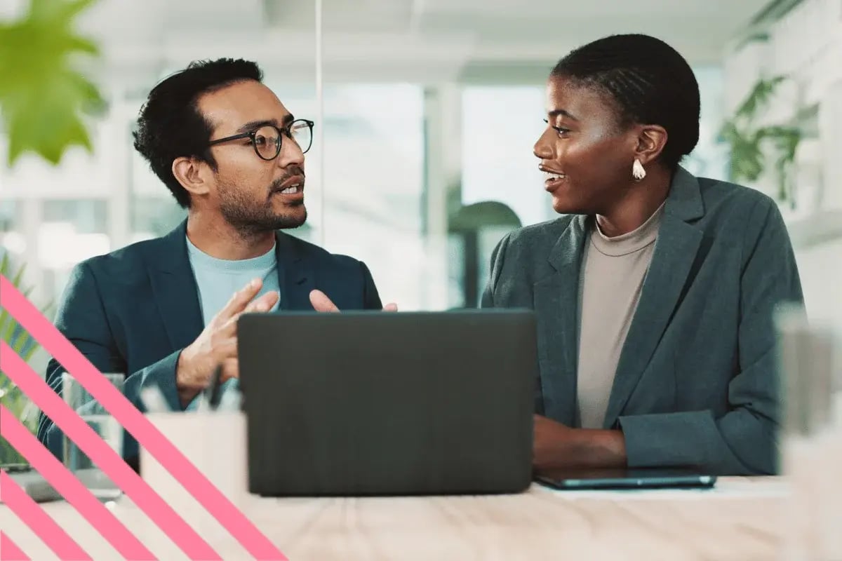 A man and a woman in business attire have a lively discussion at a table with a laptop. Bright office setting.