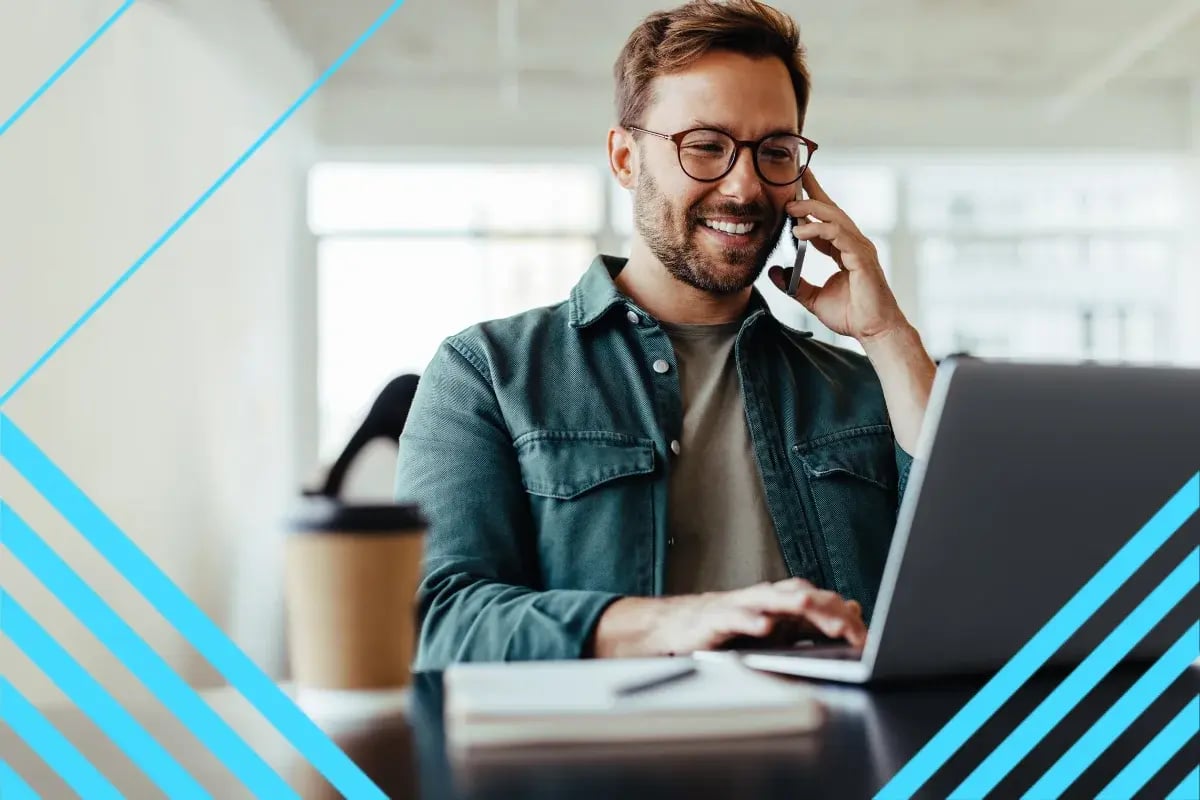Smiling bearded man in glasses works on a laptop while talking on a phone in a bright office, framed by blue geometric accents.