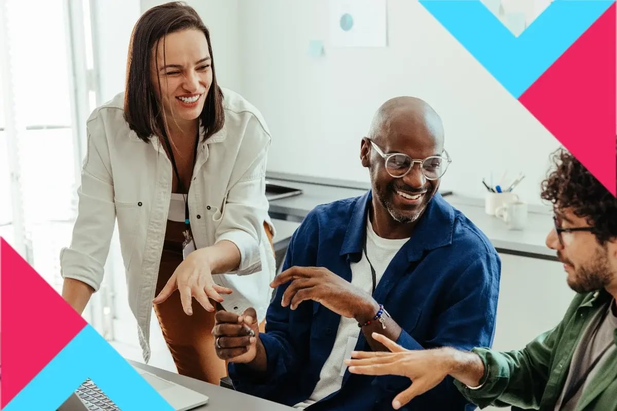Three diverse coworkers collaborate and laugh around a laptop in a bright office, conveying teamwork and enthusiasm, framed by pink and blue graphics.