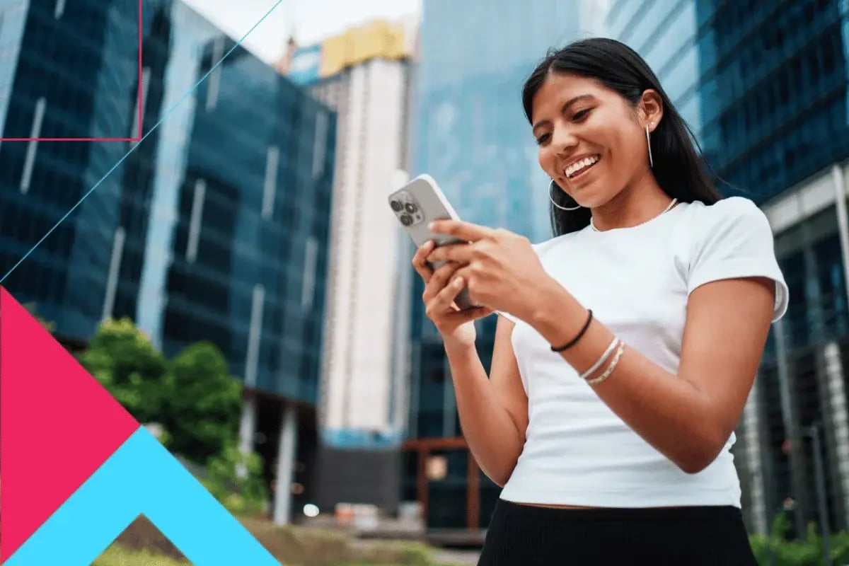 A woman smiling and looking at her phone stands outside modern glass buildings. 