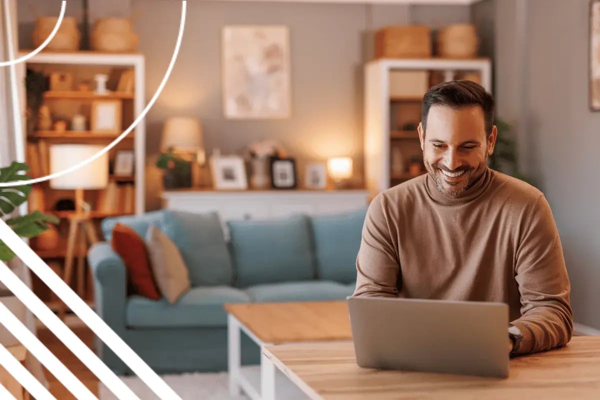 Smiling man working on a laptop at a wooden table in a cozy living room with soft lighting, a teal sofa, and shelves with decor in the background.