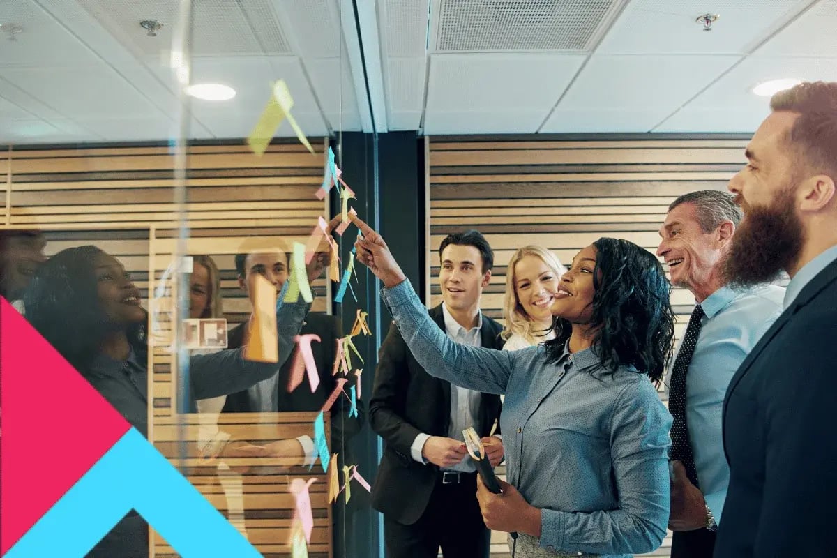 A diverse group of five professionals smiling and discussing colorful sticky notes on a glass wall in a modern office, creating a collaborative atmosphere.