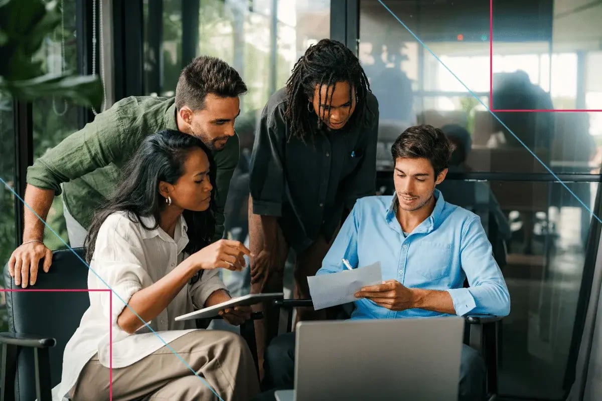A diverse team of four collaborates around a laptop, with two standing and two seated reviewing documents in an engaged discussion.
