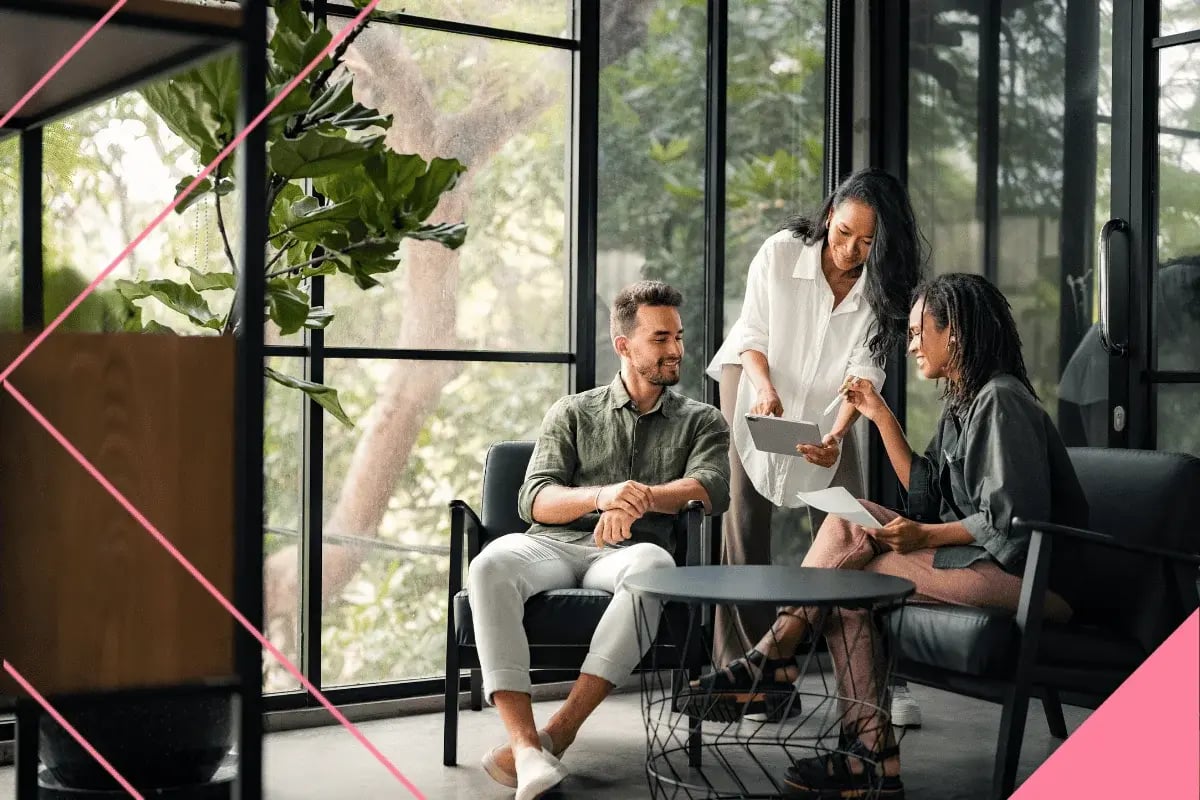 Three individuals are collaborating in a modern, sunlit office with large windows.