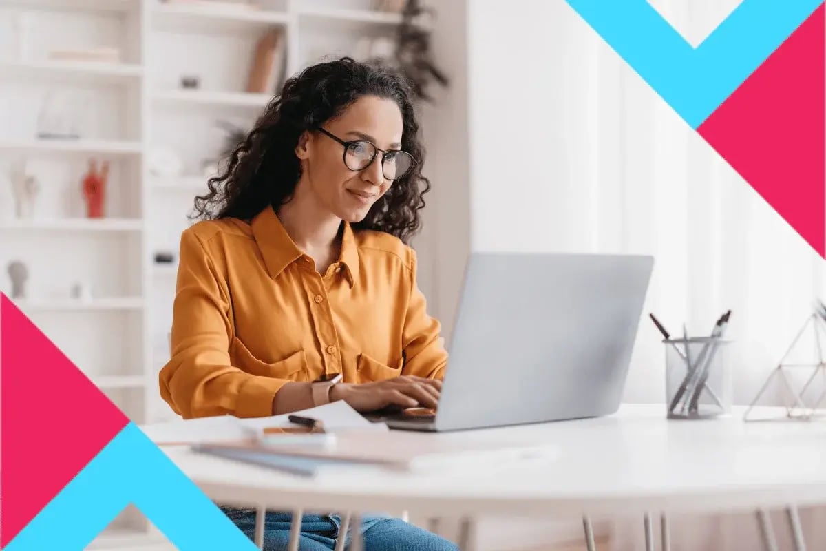 A woman with curly hair and glasses works on a laptop at a white desk. She wears a mustard-colored blouse.