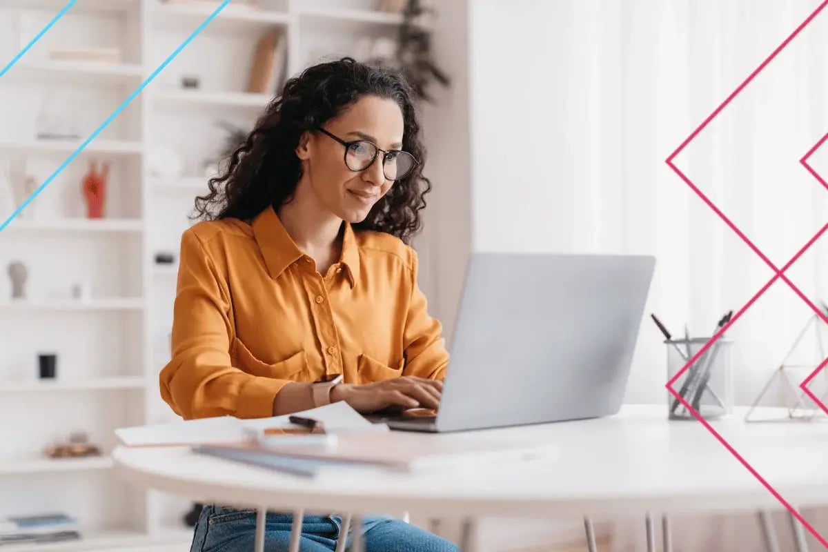Woman with curly hair and glasses using a laptop in a bright modern office with shelves.
