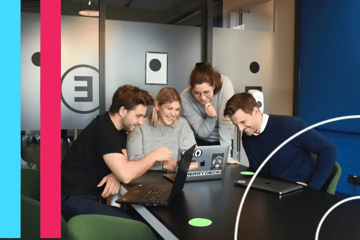 A group of four colleagues smiles and gathers around a laptop in a modern office setting, suggesting teamwork and a positive work environment.