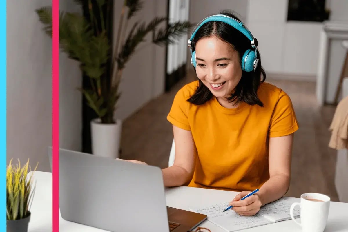 A woman in a yellow shirt and blue headphones smiles at a laptop while taking notes.