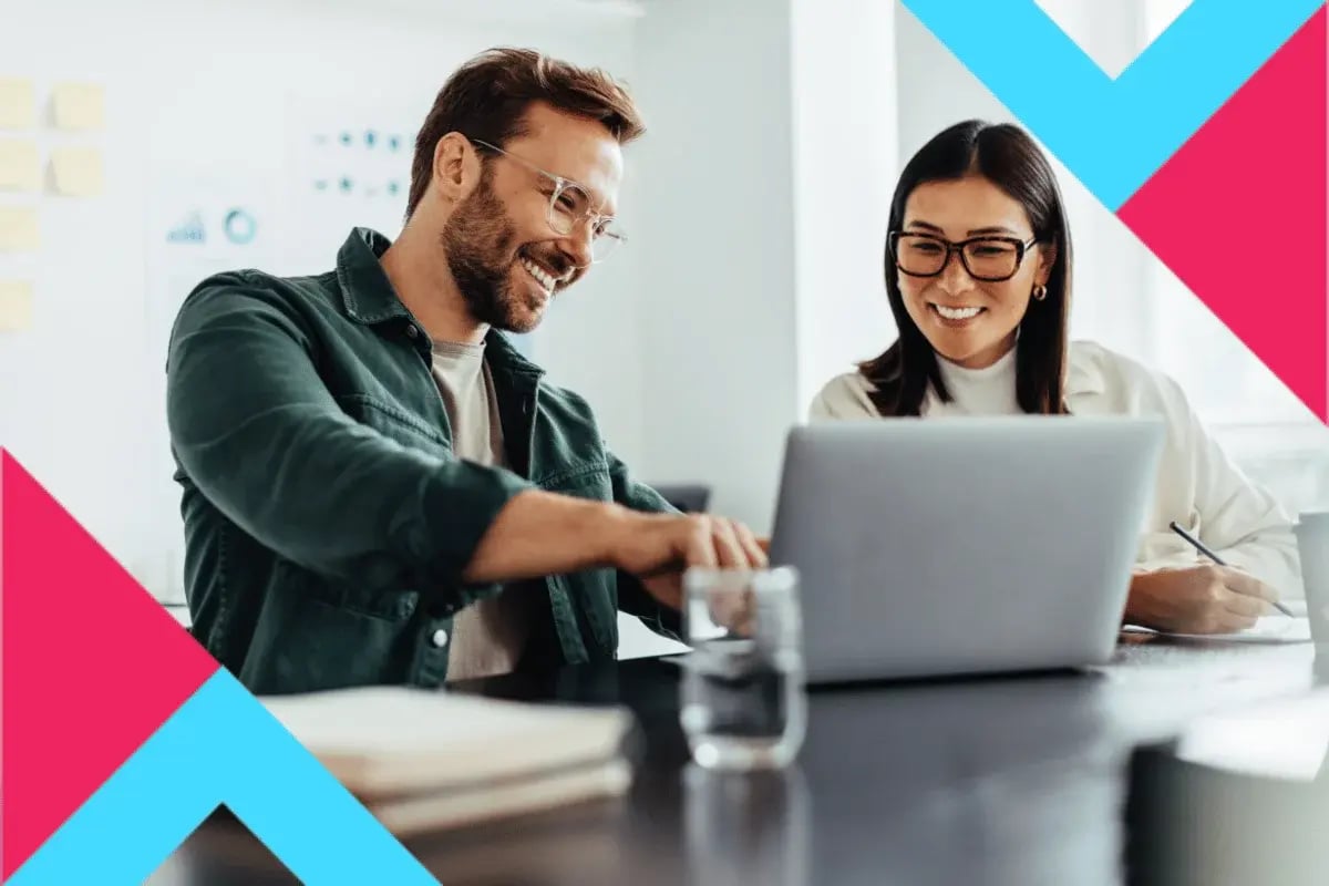 Two people smiling and collaborating at a desk, looking at a laptop.