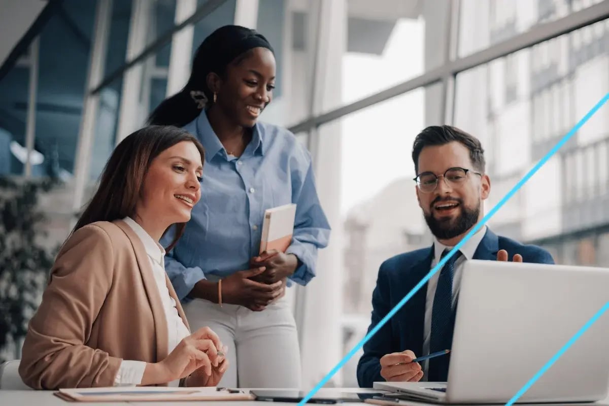 Three colleagues in business attire happily discuss around a laptop in a modern office with large windows. 