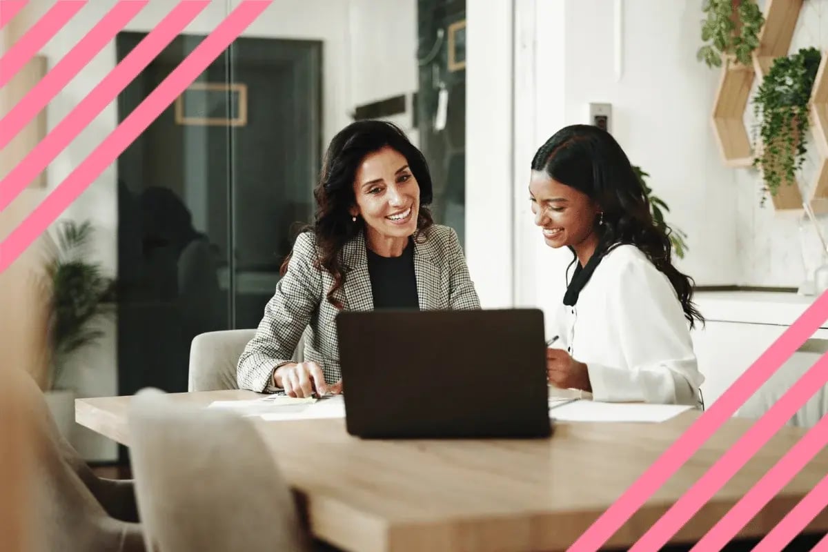 Two women smiling and discussing work at a table with a laptop. 