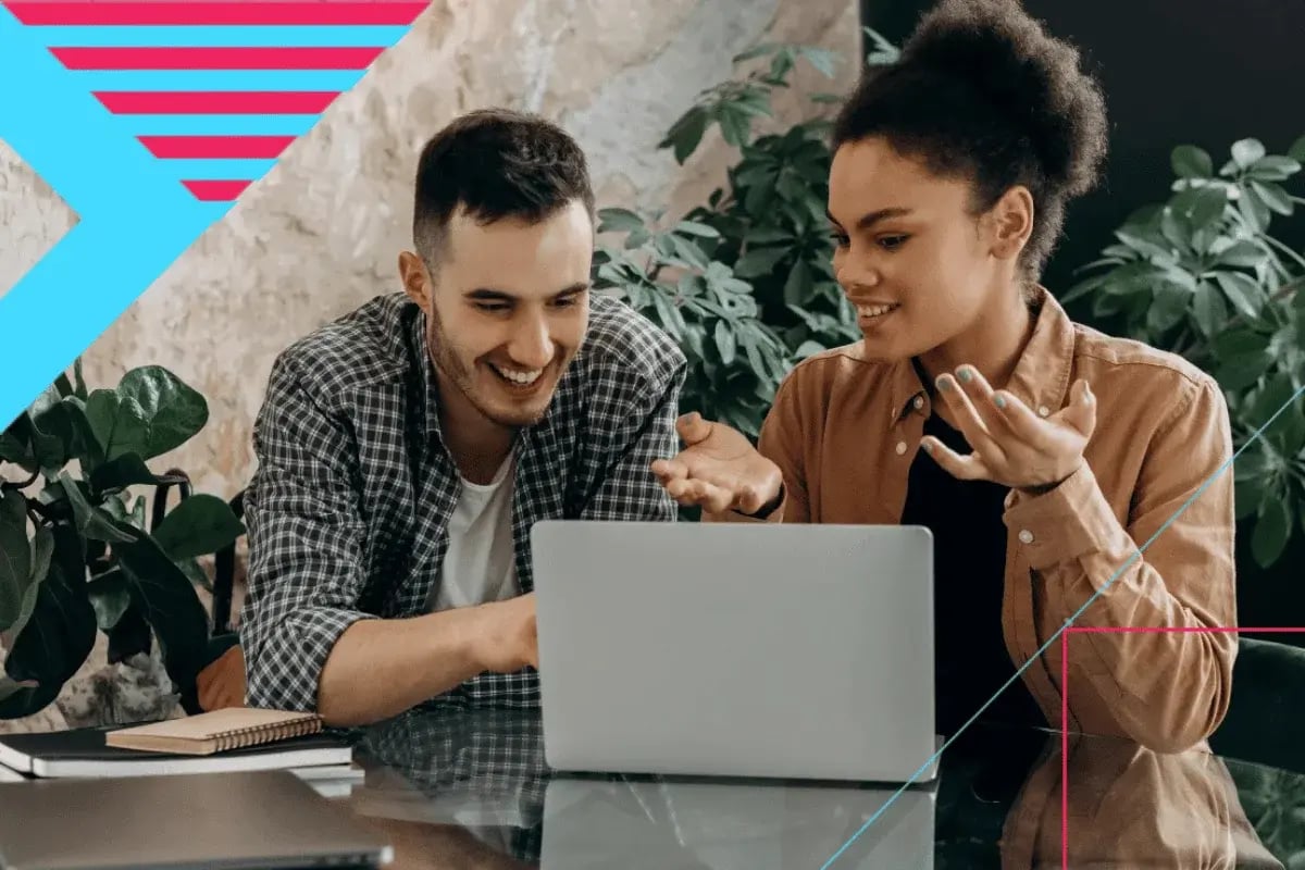 Two coworkers smiling while reviewing a project on a laptop in a modern office with indoor plants.
