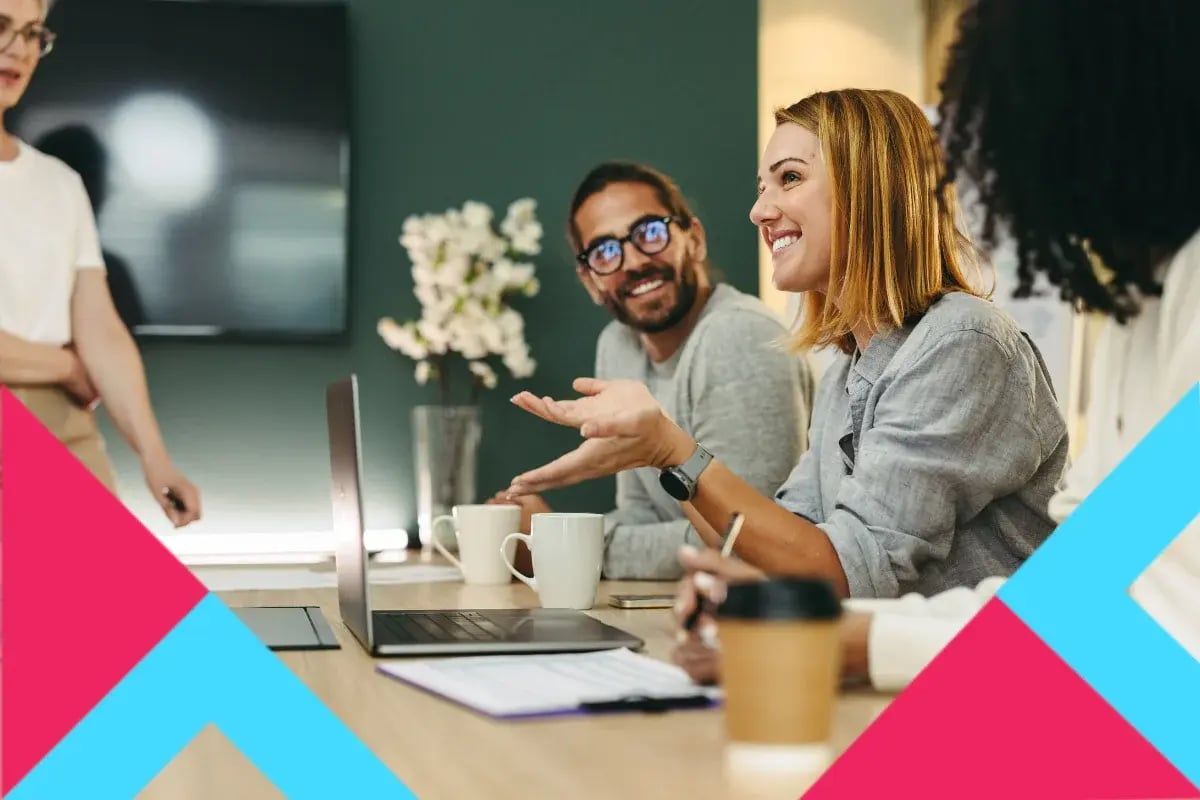 A diverse group of professionals sit around a wooden conference table, collaborating with a laptop, documents, and coffee mugs in a modern office.