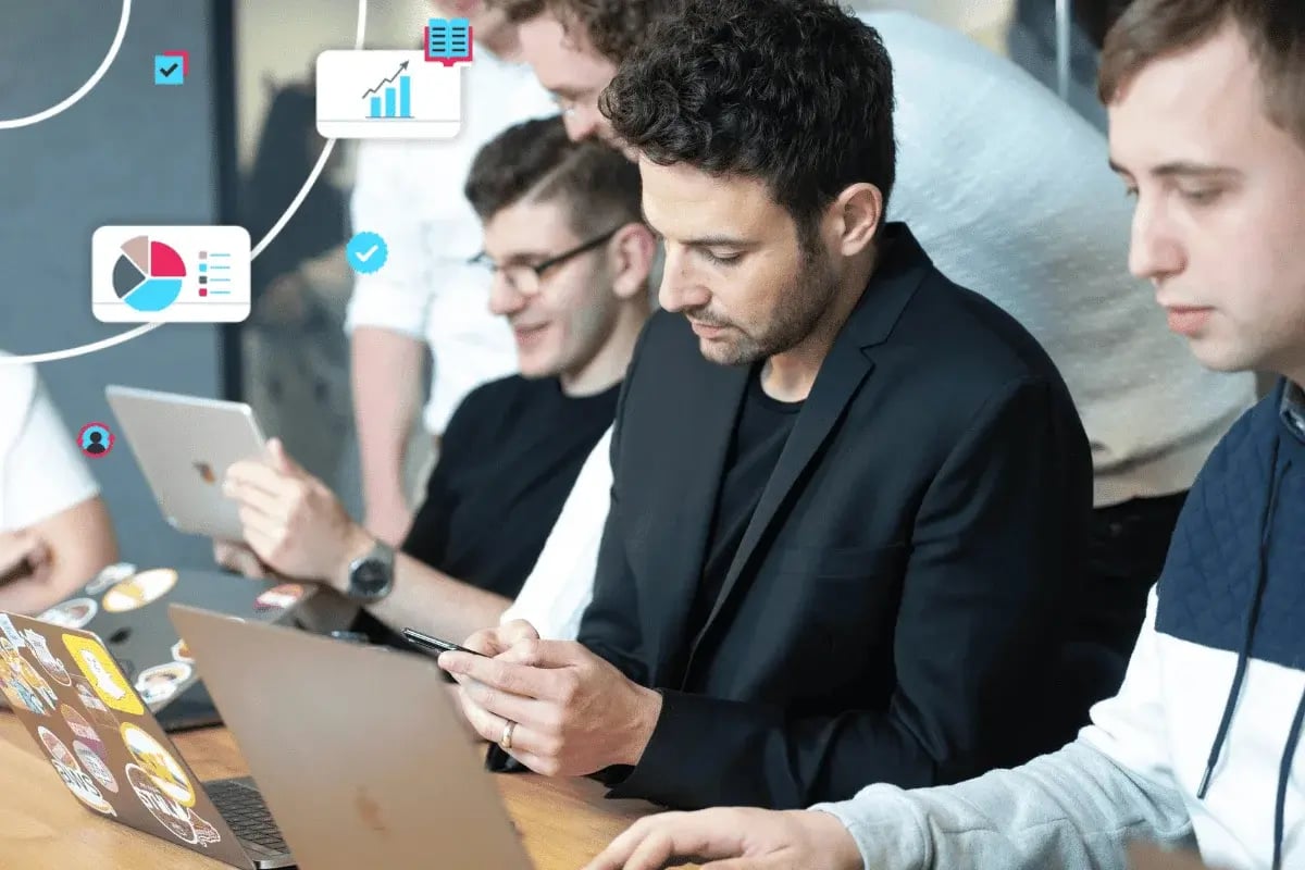A group of people sits at a table using laptops and tablets, focused and collaborative.