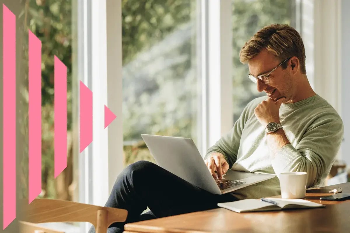 A man sits at a wooden table by a large window, focused on a laptop. He is smiling, wearing glasses, with coffee and a notebook nearby. Pink triangular graphics overlay.
