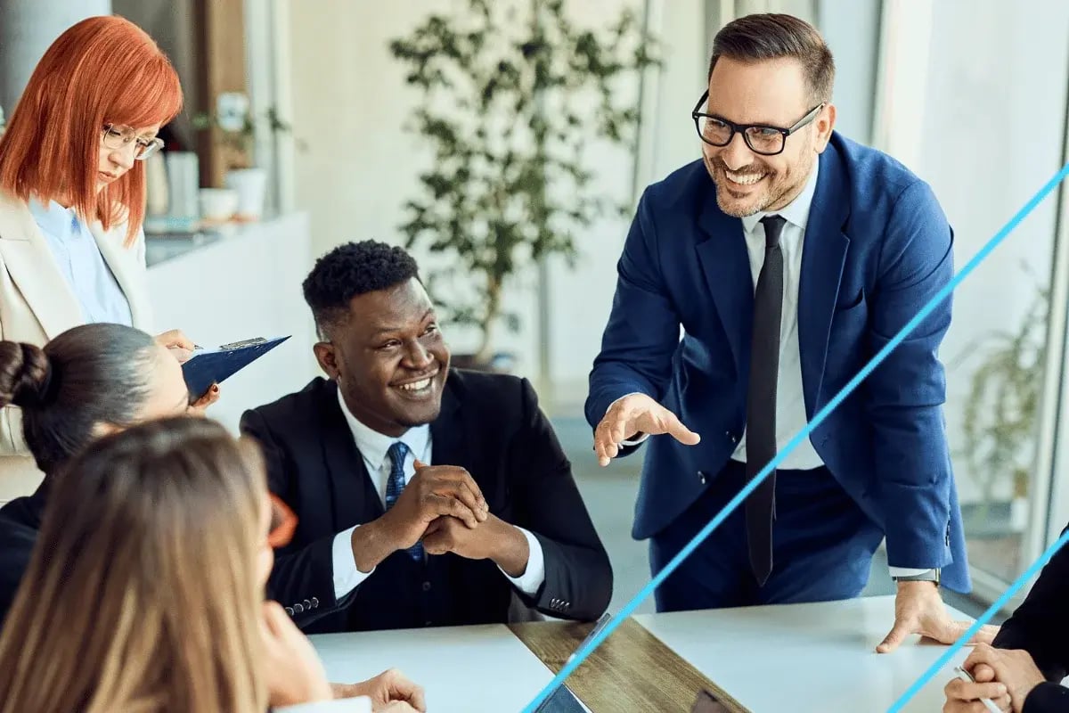 A diverse group of professionals in a meeting room. A smiling man in a blue suit leans over the table, engaging others. The atmosphere is collaborative.
