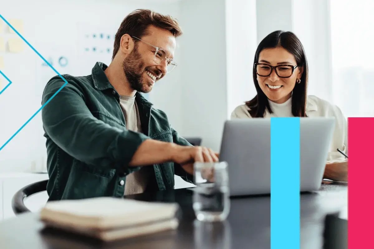 A man and a woman smile and discuss something on a laptop in a bright office. A glass of water and a notebook sit on the table, creating a collaborative mood.