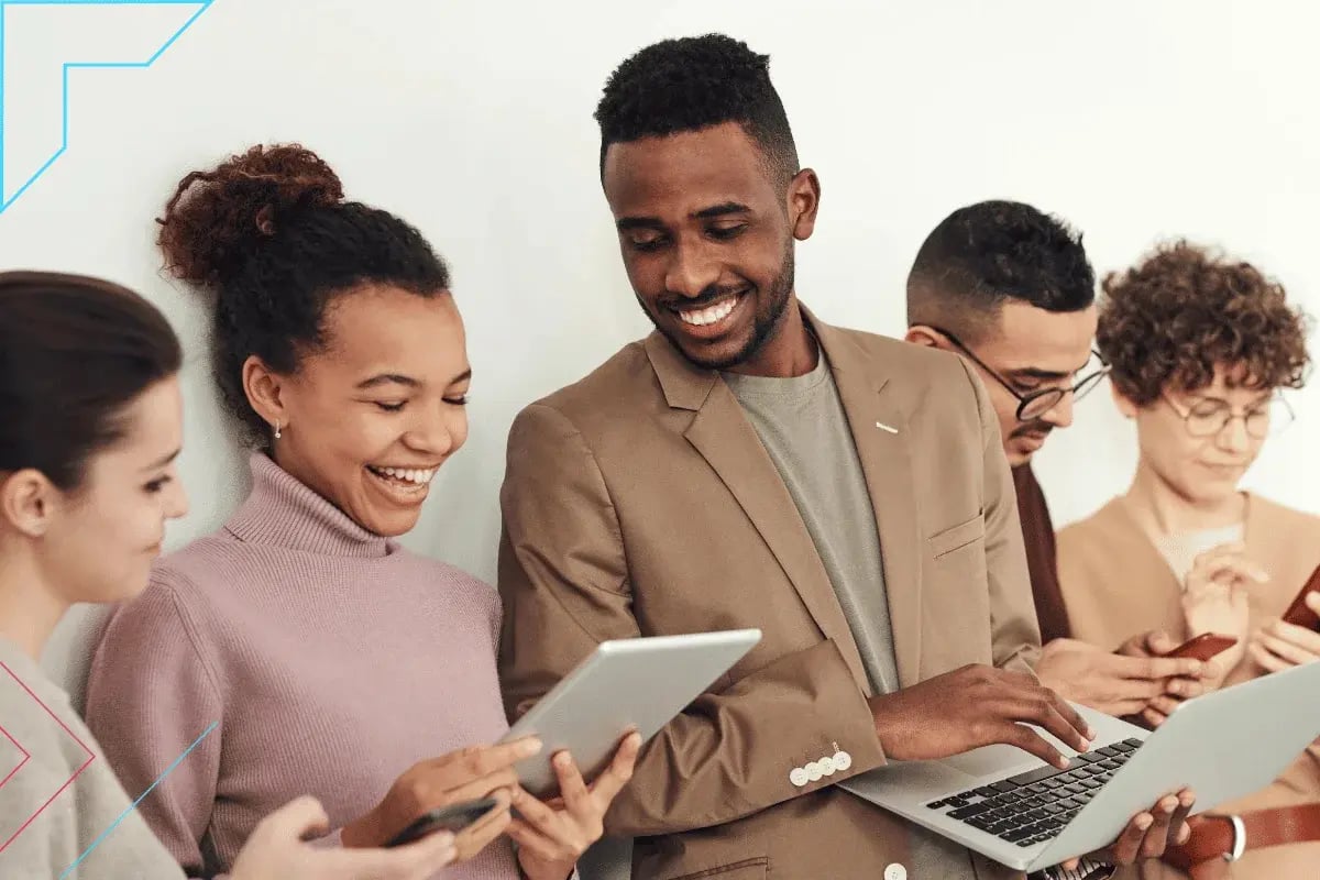 A diverse group of five people stand against a white wall, smiling and interacting with digital devices like tablets and laptops, conveying collaboration and positivity.