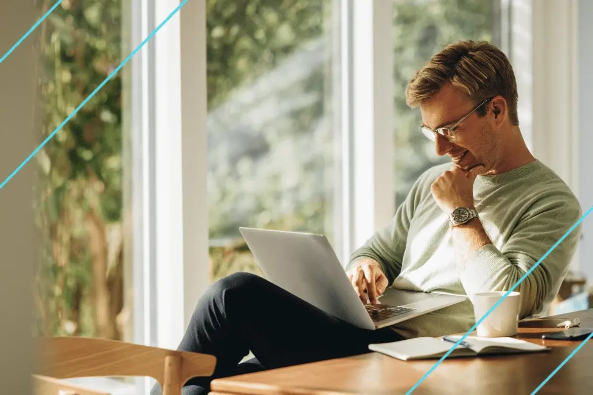 A man in glasses sits at a wooden table with a laptop, deep in thought. Sunlight streams through large windows, creating a serene and focused atmosphere.