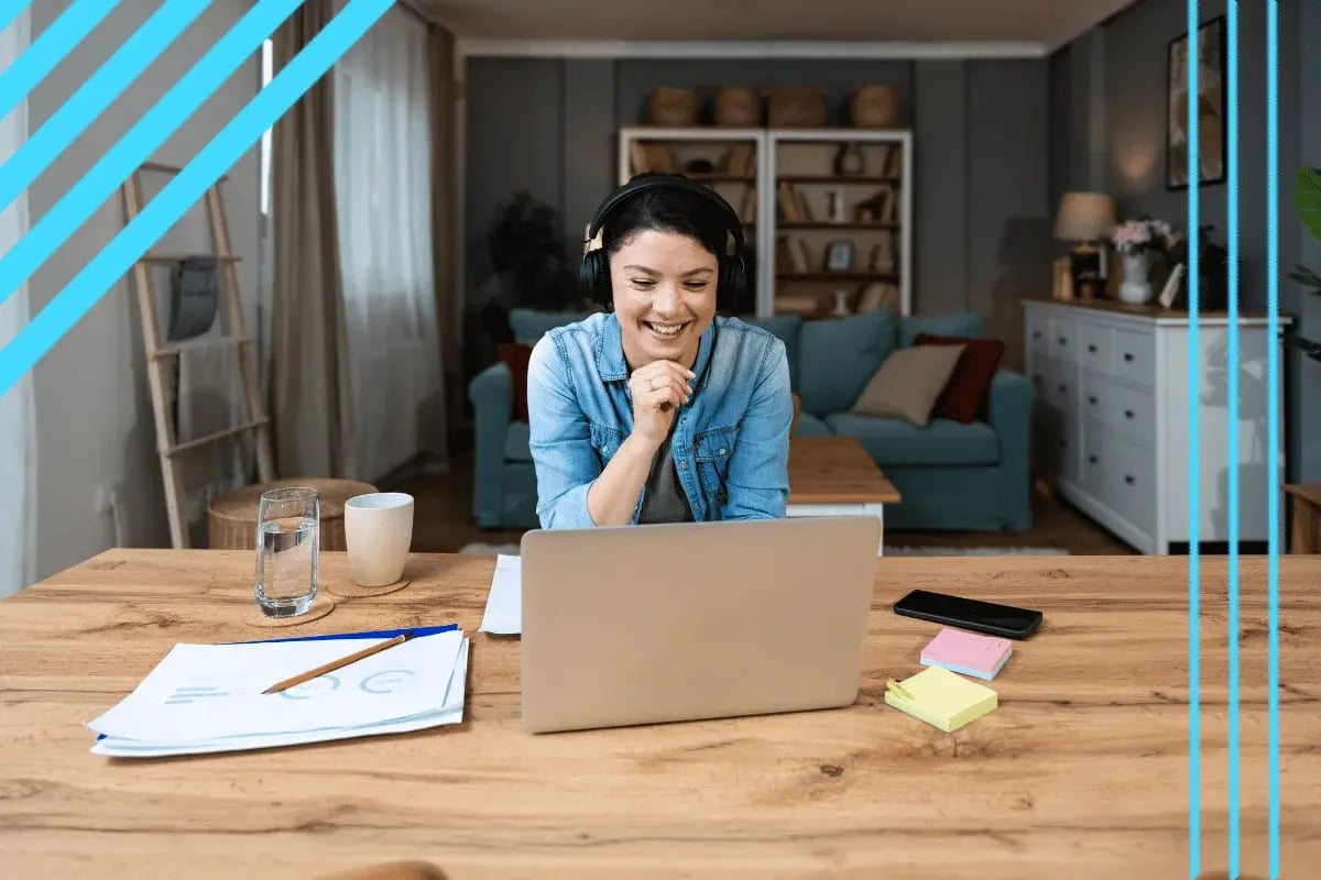 Smiling woman in denim shirt and headphones video chats on a laptop at a wooden table in a cozy living room with notes.