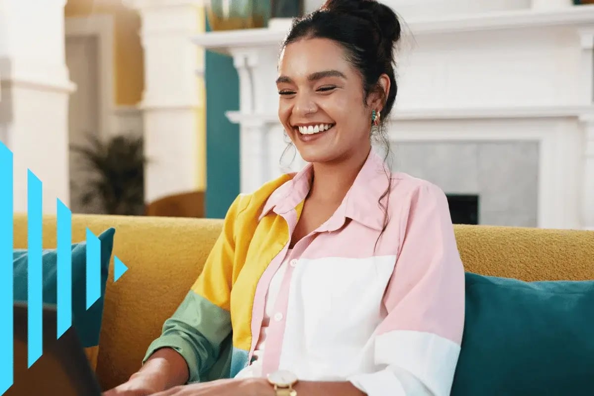 A woman with a bun hairstyle smiles warmly while sitting on a yellow couch in a cozy, stylish room.