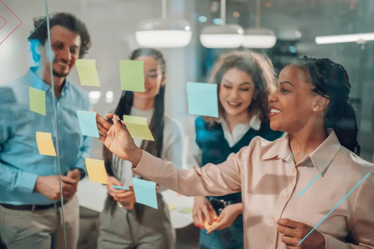 A diverse group of four people in an office smiling and discussing ideas. They are placing colorful sticky notes on a glass wall, suggesting a collaborative and creative atmosphere.