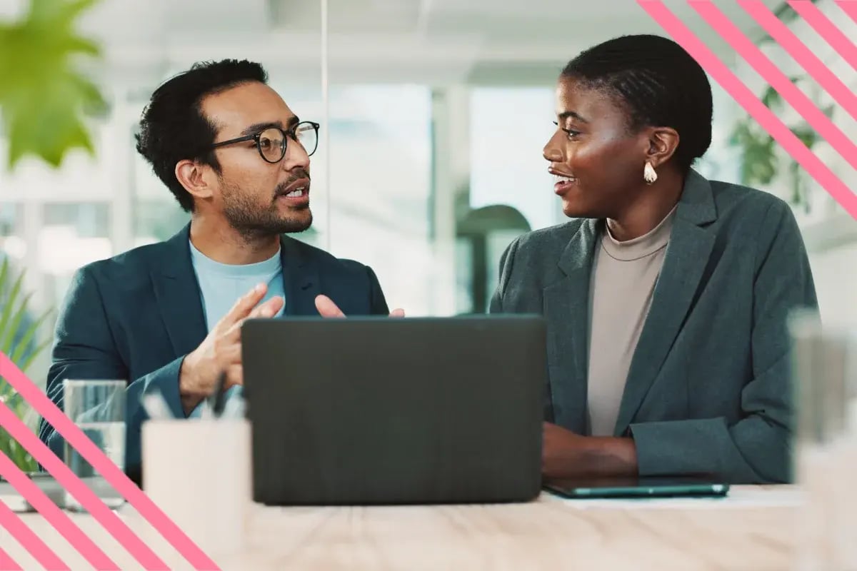 Two professionals in an office discuss ideas, smiling. A laptop is open on the table between them.