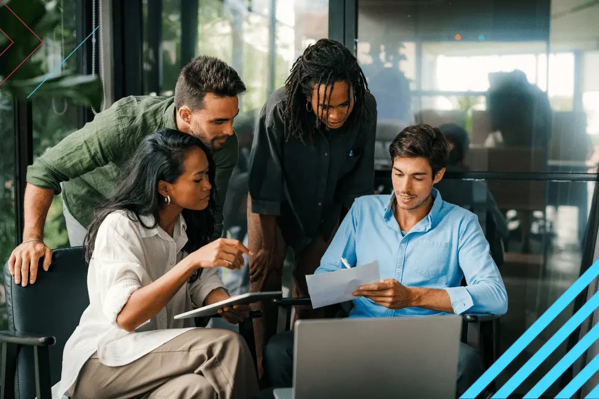 A group of four individuals collaborate around a laptop in a modern office setting, engaged in discussion and reviewing documents.