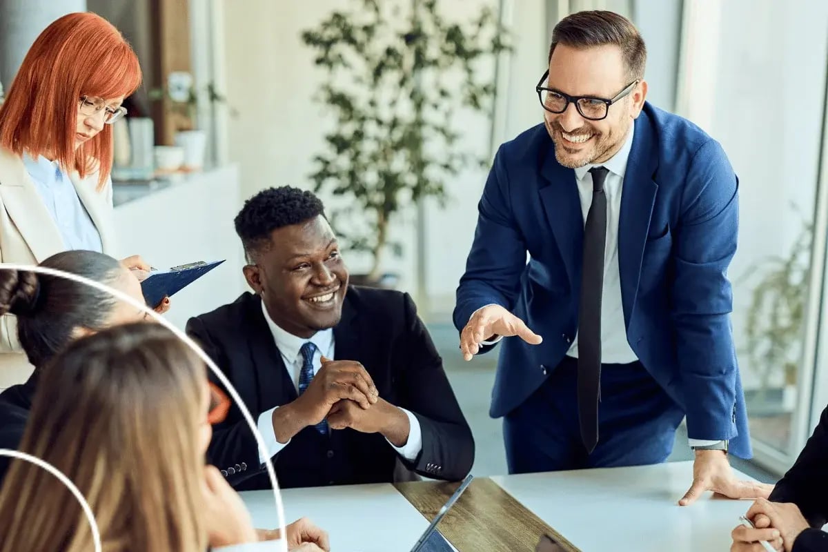 Business team in a meeting room engaged in discussion, with one man standing and presenting to seated colleagues.