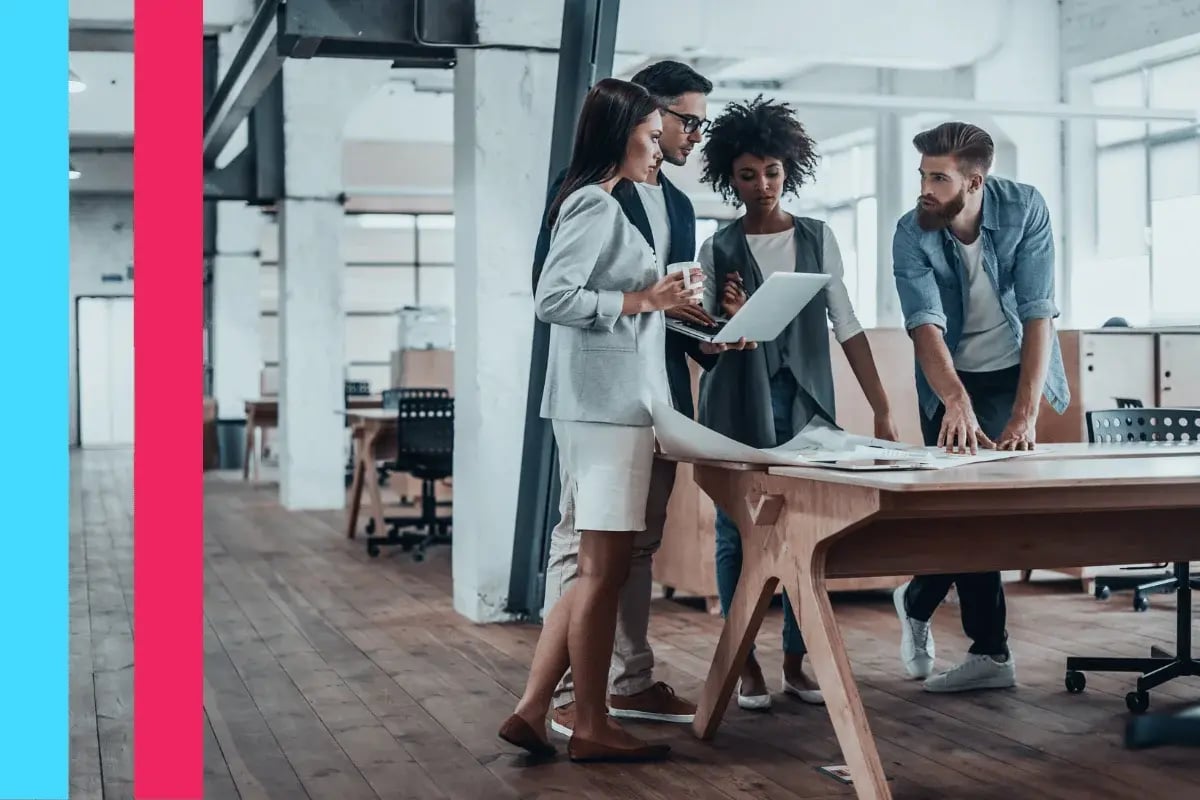 Four people in a modern office gather around a table, engaged with a laptop and documents.