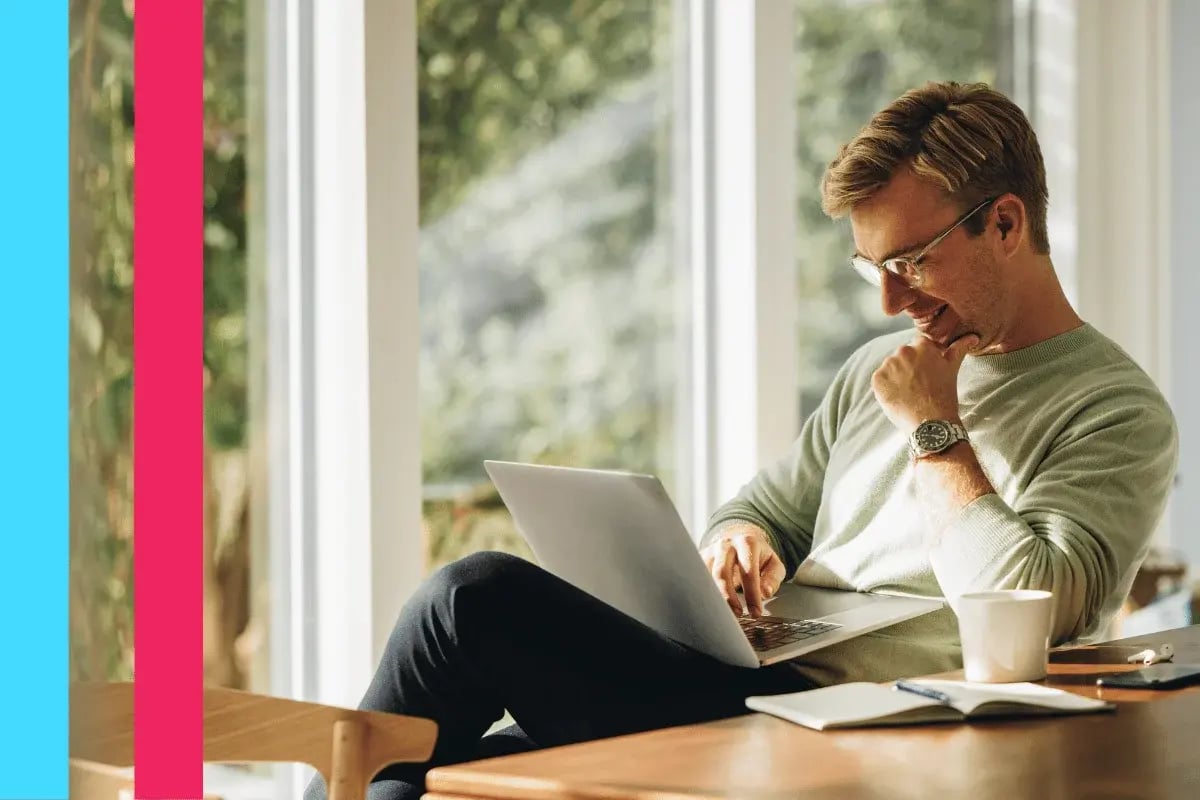 A man in glasses, wearing a light sweater, sits comfortably at a wooden table using a laptop. 
