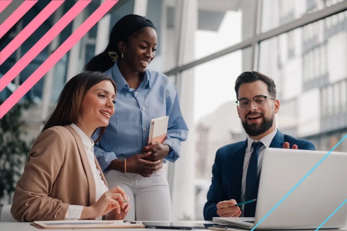 Three diverse business professionals collaborate around a laptop in a bright modern office, smiling during an upbeat team discussion.