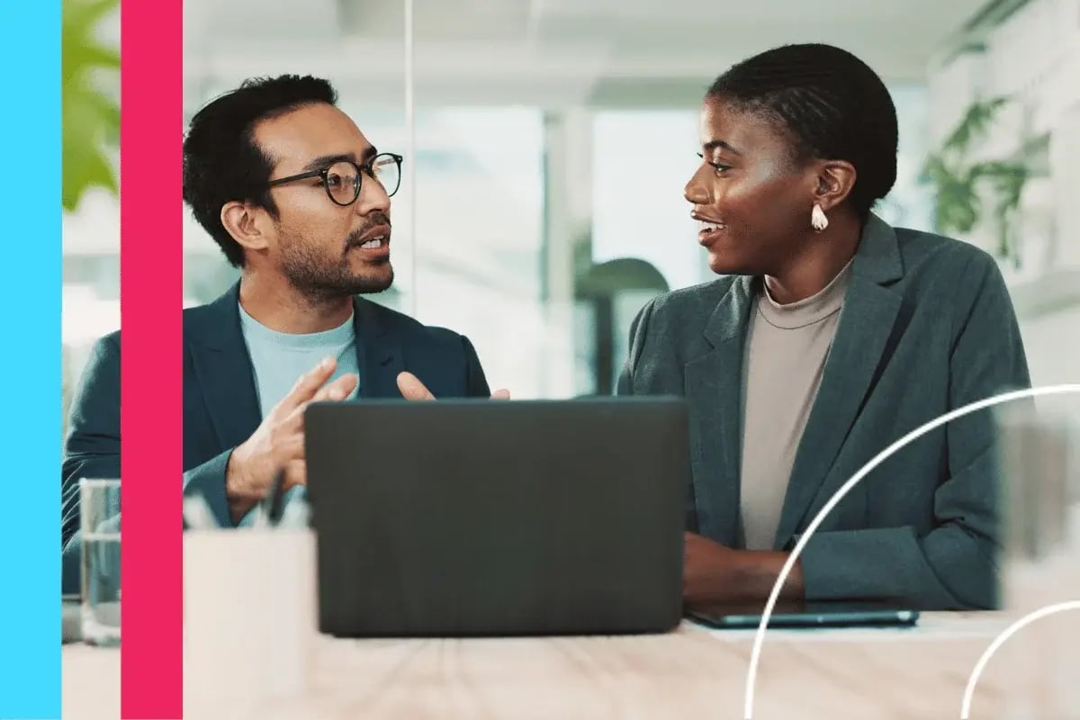 Two professionals engaged in a discussion at an office table, with an open laptop between them.