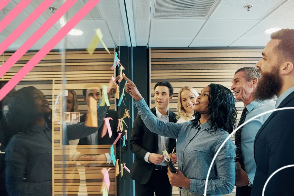 A diverse group of five people in business attire collaborates with colorful sticky notes on a glass wall, appearing engaged and smiling.