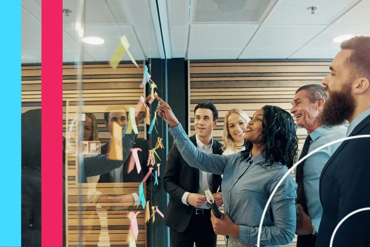 A diverse group of six professionals collaborates cheerfully around a glass wall with colorful sticky notes, suggesting a creative team environment.