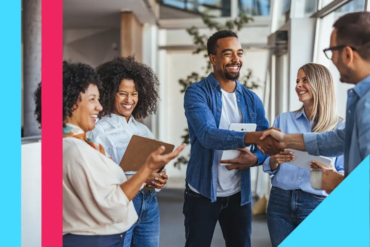 A diverse group of four professionals in casual attire share a joyful moment in an office setting, shaking hands and smiling, creating a friendly atmosphere.