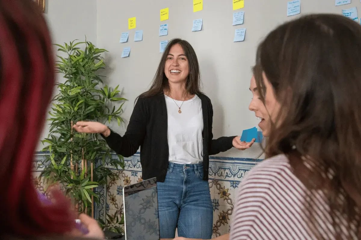 A woman gestures while speaking in a meeting, with sticky notes on the wall and participants seated in front of her.